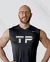 Ein lächelnder Mann mit Brille und Bart trägt ein schwarzes ärmelloses Hemd mit „PERSONAL TRAINING TP“.