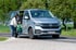 Frau hilft Kind beim Einsteigen in einen silbernen VW Transporter am Straßenrand vor grünen Bäumen.