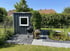 Holzschuppen mit weißem Fenster neben Grill und Teich mit Seerosen im Garten.
