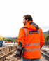 Bauarbeiter in oranger Warnjacke mit Knebel-Logo blickt auf eine Baustelle mit ausgehobenem Graben und Bagger.