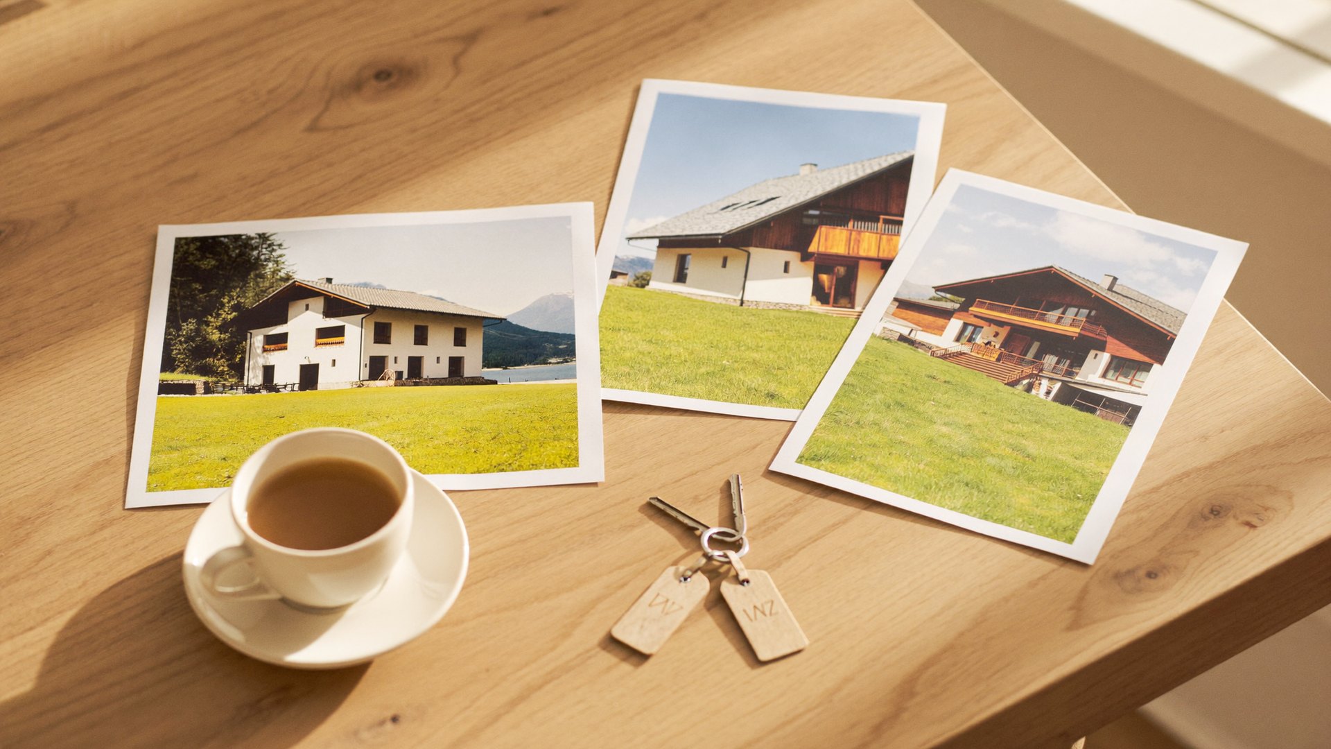 Heller Küchentisch am Morgen: Fotoabzüge von Häusern in Kärnten, zwei Schlüssel mit Holzanhängern und eine Tasse Kaffee