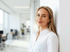 Junge blonde Frau in weißem Hemd lehnt an einer weißen Wand in einem modernen Büro.