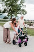 Verena und Mädchen nutzen lila Kinderrollator, begleitet von Seniorin mit Gehhilfe auf einem asphaltierten Weg im Freien