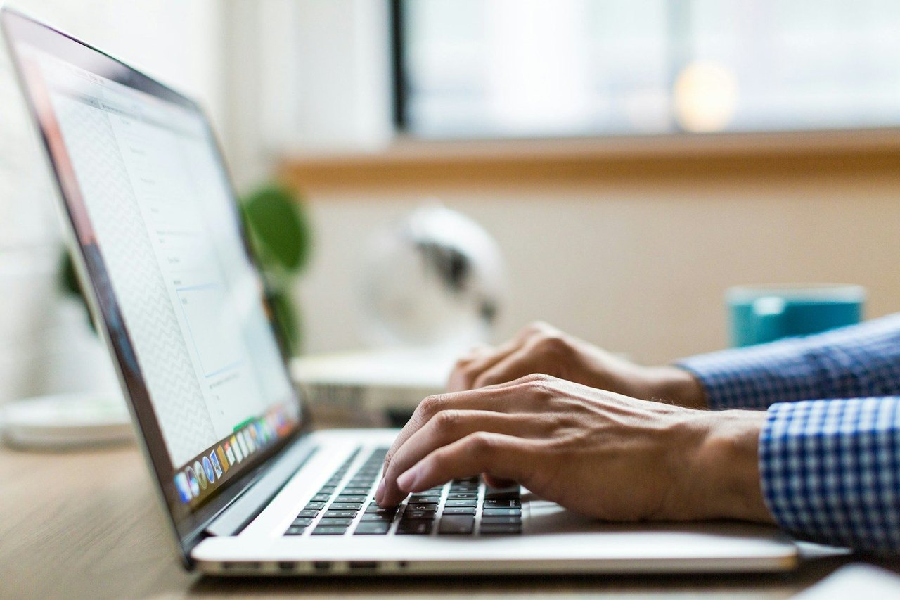 Hände tippen auf einem silbernen MacBook am Schreibtisch, im Hintergrund unscharf ein helles Büro mit Fenster