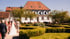 Ein Brautpaar und Hochzeitsgäste gehen durch einen Garten auf ein historisches Gutshaus mit Walmdach zu.