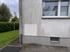 Weiße Hauswand mit einem frisch gestrichenen, rechteckigen Bereich unter einem Fenster, daneben ein Regenfallrohr und grüner Garten.