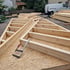 Zwei hölzerne Dachsparrenrahmen mit Zwingen auf einer OSB-Platte, ein Lieferwagen und Holzstapel im Hintergrund.
