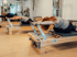 Frauen trainieren auf hölzernen Pilates-Reformern in einem hellen Studio und führen Dehnübungen nach vorne aus.