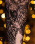 Detailliertes Schwarz-Grau-Tattoo einer Eule mit Herz-Ausschnitt im Gefieder vor goldenem Bokeh-Hintergrund.