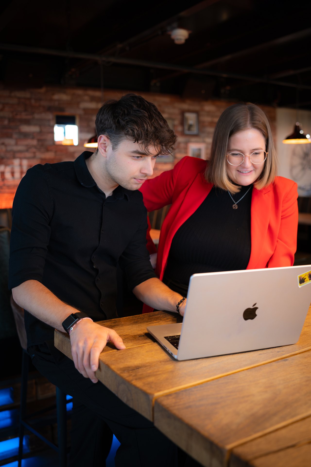 Zwei Personen blicken auf einen Laptop, ein Mann im schwarzen Hemd und eine Frau in einer roten Jacke.