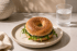 Sesam-Sauerteig-Bagel mit Frischkäse und Rucola auf einem Teller neben einem Wasserglas.
