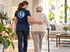 Eine Pflegekraft stützt eine Seniorin mit Gehstock beim Gehen durch ein helles Wohnzimmer.