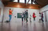 Ein Mann und sechs Kinder springen in einem Turnsaal mit Holzboden und weißen Wänden.