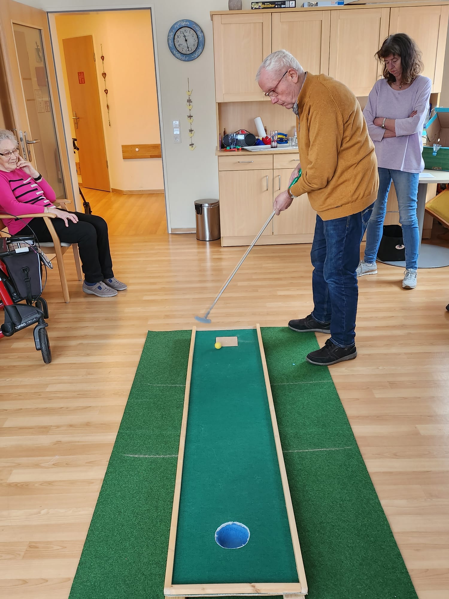 Ein Mann spielt Minigolf auf einer grünen Bahn in einem Gruppenraum.