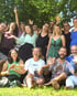 Eine große Gruppe lachender Menschen posiert fröhlich auf einer grünen Wiese vor Bäumen im Freien.