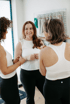 Drei lächelnde Frauen in weißer Sportkleidung und schwarzen Leggings unterhalten sich in einem hellen Fitnessstudio.