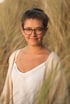 Frau mit Brille und hochgesteckten Haaren lächelt in die Kamera, umgeben von trockenem Gras im Sonnenlicht.