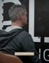 Ein Mann mit Brille und grauem Kapuzenpullover sitzt vor einer Wand mit großen Buchstaben und dem Text „nicht empfehlen“.
