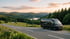Schwarzer Campervan am Straßenrand mit Blick auf bewaldete Hügel, einen See und Sonnenuntergang.