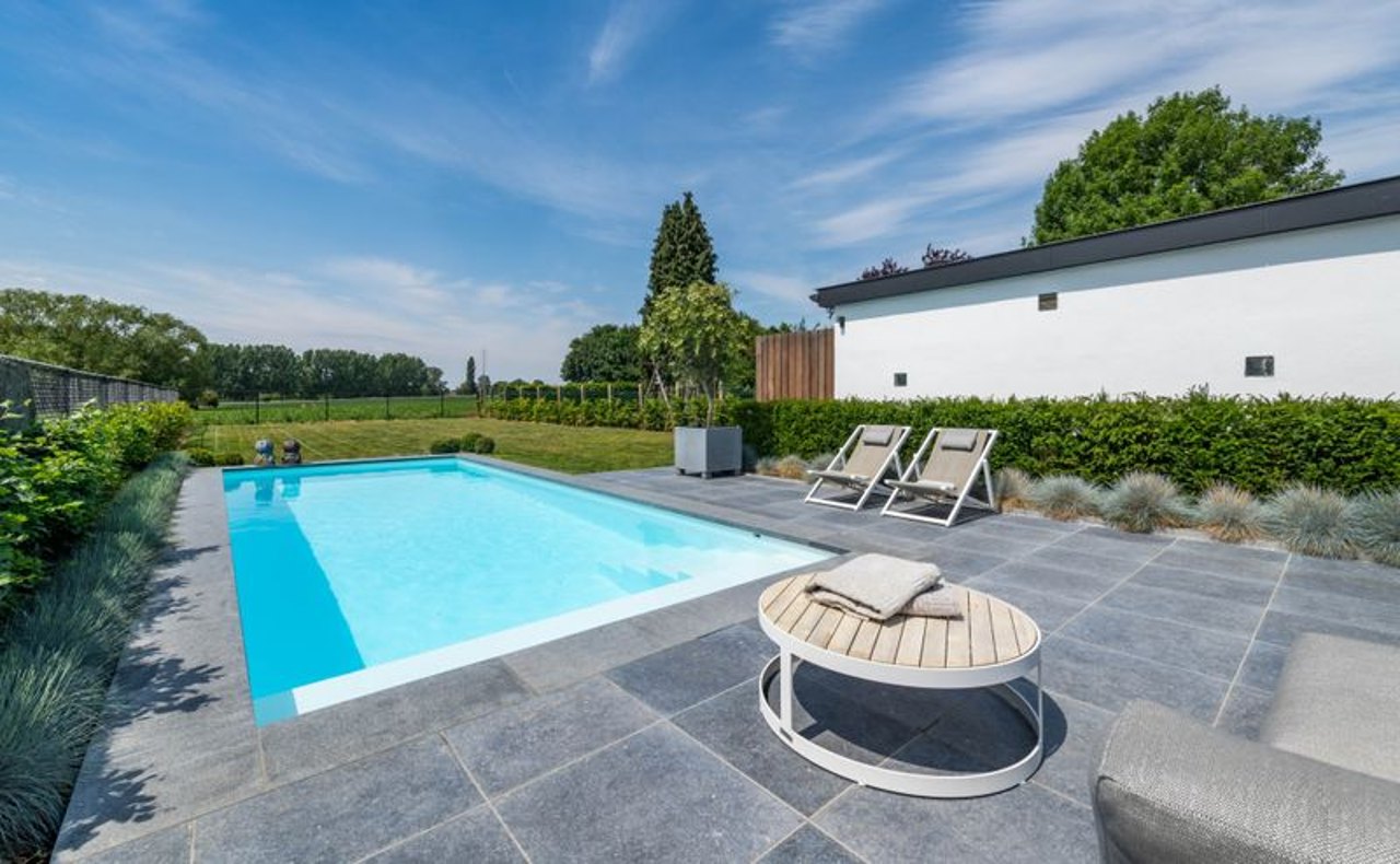 Rechteckiger Pool mit blauer Fliesenverkleidung, umgeben von dunklem Pflaster und Liegestühlen, gebaut von Calis Gartenbau