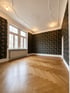 Leerer Altbau-Raum mit Fischgrätparkett, dunkler Blumentapete und verzierter Stuckdecke mit Deckenleuchten.