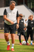 Ein Fußballtrainer in grauem T-Shirt mit "TSA SINCE 2018" und schwarzen Shorts auf einem Rasenplatz mit Kindern.