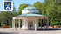 Weißer Rundtempel mit grünem Kuppeldach und Säulengang im Kurpark Bad Oeynhausen vor grünen Bäumen mit Stadtwappen.