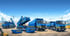 Mehrere blaue LKW und Kipper auf einer Baustelle mit klarem blauem Himmel und Kränen im Hintergrund.