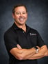 Smiling man in a black polo shirt with a chiropractic logo, arms crossed, standing against a dark gray background.