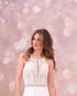 Frau mit braunem Haar trägt ein weißes Brautkleid aus Spitze mit Neckholder-Ausschnitt vor einem rosa Bokeh-Hintergrund.