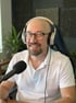 Ein Mann, Michael Beranek mit Glatze, Bart, Brille und weißen Kopfhörern sitzt lächelnd vor einem Mikrofon in einem Büro. Er trägt ein weißes Poloshirt.