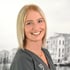 Eine lächelnde junge Frau mit blondem Bob und dunklem Kittel vor einer verschwommenen Schwarz-Weiß-Kulisse.