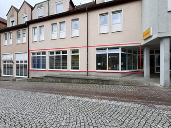 Fassade des Gebäudes mit dem markierten Ladenlokal im Erdgeschoss, davor ein gepflasterter Platz