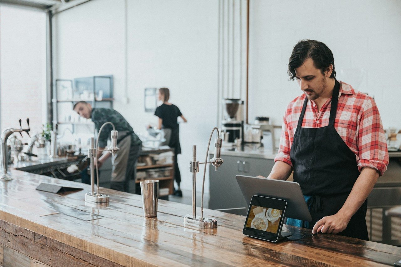 Barista mit Schürze bedient ein Tablet-Kassensystem an einer Holztheke im Café, im Hintergrund arbeiten zwei Kollegen