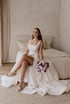 Lächelnde Braut im weißen Hochzeitskleid mit Beinschlitz sitzt auf einem Bett und hält einen lila-rosa Blumenstrauß.