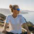 Blonde Frau in weißem T-Shirt und blauer Sonnenbrille auf einem Fahrrad blickt nach rechts, Küste im Hintergrund.