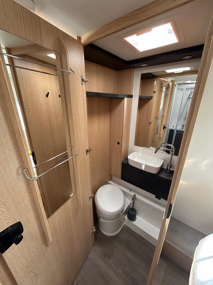 Helles Badezimmer in einem Reisemobil mit Holzwänden, weißer Toilette, Waschbecken und Spiegel.