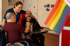 Eine lächelnde Frau in blauer Uniform steht hinter zwei lachenden Männern in Rollstühlen, Regenbogenflagge im Vordergrund.