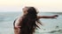 Woman with long brown hair, wearing a black top, arms outstretched, head tilted back, on a sandy beach.