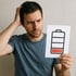 Ein besorgter Mann hält ein Blatt Papier mit einem Batteriesymbol und niedrigem Ladestand, welche sine geringe, gesundheitliche Energie widerspiegelt. 