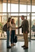 A male teacher in a blazer shakes hands with a female student and her mother in a school lobby.