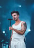 Sänger in einem weißen Tanktop mit tätowierten Armen, singt in ein Mikrofon vor einem Hintergrund aus blauem Bühnenlicht und Live-Atmosphäre.