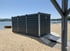 Dunkelgrauer, rechteckiger Container mit zwei Metallrampen auf einem Sandstrand am Seeufer.