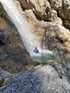 Eine Person in blauer Ausrüstung steht unter einem Wasserfall in einem Felsbecken.