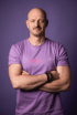 A bald man with a mustache, wearing a purple t-shirt with "müller" written in pink, stands with arms crossed.