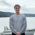 Ein lächelnder junger Mann mit grauem Pullover steht vor einem See mit Boot und bewaldeten Hügeln im Hintergrund.