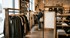 Woman browsing clothes in a retail store with wooden display racks and a blank white sign.