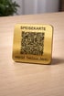 Goldene Speisekarte mit QR-Code und Text "SPEISEKARTE IMBISS THESSALONIKI" auf einem Holztisch.