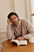 Ein Mann mit Bart und braunem Strickpullover sitzt an einem Holztisch vor einem aufgeschlagenen Buch und einer Tasse.