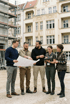 Fünf Bauarbeiter, drei Männer und zwei Frauen, besprechen Baupläne auf einer Baustelle mit Gebäuden im Hintergrund.
